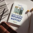 A képen a Hogyan neveljünk boldog kutyát című könyv látható a háttérben egy kutyáját ölelő nő.
