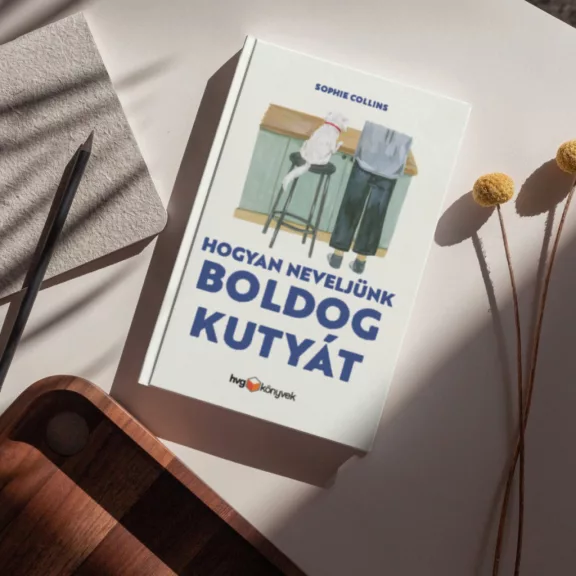 A képen a Hogyan neveljünk boldog kutyát című könyv látható a háttérben egy kutyáját ölelő nő.