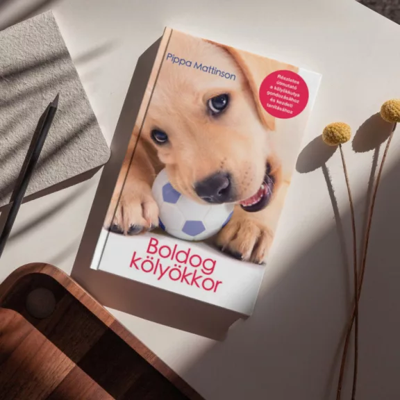 A képen a Boldog kölyökkor című könyv látható a háttérben egy labrador kölyök.