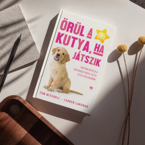 A képen az Örül a kutya ha játszik című könyv látható a háttérben egy kiskutya.