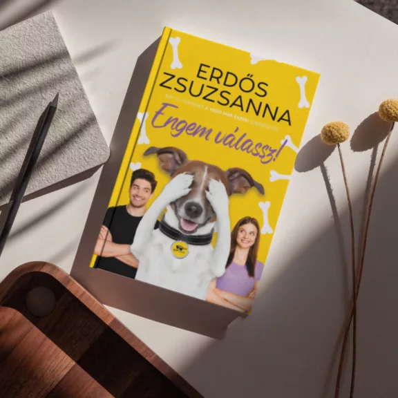 A képen az Engem válassz című könyv látható a háttérben egy kutya.