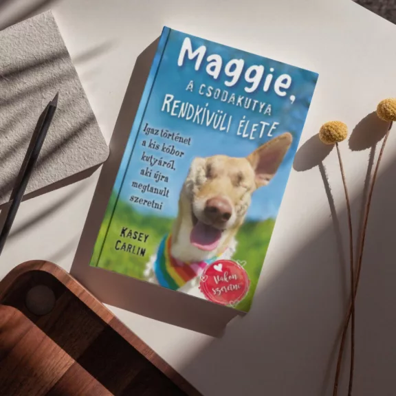 A képen a Maggie a csodakutya rendkívüli élete című könyv látható a háttérben egy réttel.