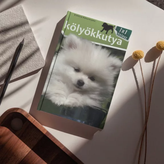 A képen a Kölyökkutya 1x1 kalauz című könyv látható a háttérben egy pomerániai törpespicc.